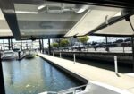 View from under a covered dock of a marina: water canal with moored boats and a walkway lined by a railing in the background.