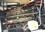 Close-up of a boat engine in a dirty engine bay, with red battery cables, black hoses, and various metal brackets visible.