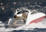White motor yacht speeding through choppy water with passengers on the deck and a hilly coastline in the background.
