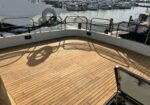 Yacht deck with wooden planks and safety netting along the railing; a marina with moored boats in the background; bicycle shadow on the deck.