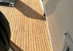 Wooden boat deck with a hatch open on the left, casting shadows over light brown planks near the water.