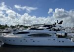 Large white luxury yacht docked at a sunny marina with other boats and palm trees in the background.