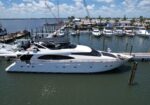 Large white luxury motor yacht docked at a marina with other boats and calm water under a blue sky with scattered clouds