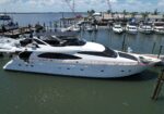 Large white luxury motor yacht docked at a marina with other boats and docks in the background under a partly cloudy sky.