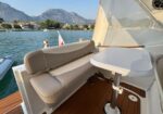 Boat deck seating with a beige cushioned bench and a white round table, on calm water with a coastal town and mountains in the background.