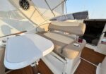 Cockpit seating area on a luxury boat with a curved beige bench around a white folding table; a red-topped cup in the cup holder, marina in the background.