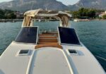 Front view from the bow of a white motorboat on calm blue water, with a beige canopy and wooden deck, mountains in the background.