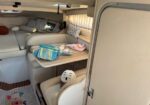 Interior of a small camper van: U-shaped sofa with cushions, a wooden table, and bags on the table; a white helmet rests on a nearby seat.