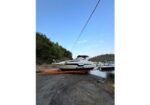 White motor yacht on an orange trailer near a rocky shore, with trees and clear blue sky in the background.