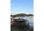White motor yacht docked at a lakeside with a person on the dock, grassy foreground and hills in the background.