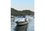 White motor yacht anchored in a calm marina with sailboats and trees in the background, bow facing the viewer.