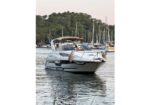 White motor yacht with a brown canopy on calm harbor water, with sailboats and trees in the background.