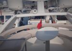Sunlit cockpit of a white luxury yacht with curved beige seating and a small white table, a red flag, and a marina full of boats in the background.