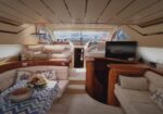 Luxurious yacht lounge with cream leather seating, wooden cabinetry, a table with flowers, and a flat-screen TV, forward helm area visible.