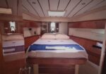 Queen-size bed in a yacht cabin with white linens, blue throw, and wooden built-in cabinetry on both sides, mirrored walls nearby.