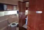 Narrow yacht bathroom with toilet and sink on the left, wooden cabinets and a door, and a glimpse of a cabin bed through a doorway.