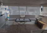 Sighted yacht cockpit lounge with a central glass table and white cushioned seating on a teak deck under a canopy, suggesting a relaxed outdoor dining area on a boat.