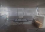 Yacht cockpit lounge with a beige sofa, glass-top table, folding chair, and a wicker basket on teak decking.
