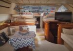 Luxurious boat cabin with cream upholstery, wooden cabinetry, a blue zigzag tablecloth on the dinette, pillows, and a flat-screen TV on the right.