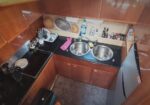 Compact kitchen with black countertop, a double sink, wooden cabinets, a stove on the left, and a small fridge on the right, with everyday items on the counter.