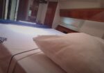 Bedroom cabin on a boat with a large white bed, a pillow in the foreground, blue throw, and wooden cabinets along the wall.