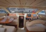 Luxurious yacht cockpit with cream leather seating, a wooden table, captain's chair, and navigation panel under a large forward windshield.