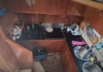 Compact boat galley kitchen with stovetop, sink, and small appliances on dark countertops amid wooden cabinets nearby trash bin and rug.