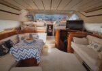 Spacious yacht salon with beige upholstered seating, a central table covered by a blue chevron cloth, decorative pillows, and a wall-mounted TV on wooden cabinetry.