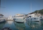 Marina with several white yachts moored in calm blue water, a clear sky overhead and a hillside town in the background.