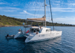 White catamaran sailing yacht anchored near a rocky coastline, with a small dinghy attached on the stern.