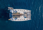 Top-down view of a white catamaran yacht on deep blue water, with solar panels and deck seating visible at the stern.