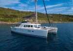 White catamaran yacht anchored near a rocky coastline under a blue sky, with sails furled and deck equipment visible
