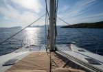 View from the bow of a sailboat looking toward the horizon with the mast and rigging in the center, sparkling sea and distant islands.