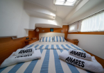 Boat cabin bedroom with a double bed in blue-and-white stripes and two rolled towels reading 'ANARES' placed at the foot.