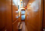 Narrow wood-paneled hallway inside a boat cabin, opening to a bed with blue-and-white striped sheets at the far end.