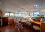 Sunlit yacht salon with a U‑shaped beige sofa, wooden dining table set for a meal, and large windows with curtains.