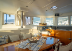 Sunlit yacht cabin with a wooden dining table, cushioned seating, and a bowl of snacks, large windows showing a coastal town.