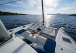 View from the deck of a white sailboat: blue cushions, striped sun loungers, and a small table with fruit, overlooking calm blue sea and distant coastline at sunset.