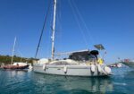 White sailboat anchored in calm turquoise water with fenders along its hull and a blue clear sky above; another boat is in the background left.