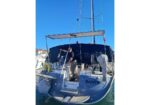 Man standing on the deck of a blue sailing yacht named SANTE in a marina, under a dark canopy with rigging around him.
