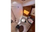 Compact boat head: white toilet beside a dark countertop sink, wooden cabinet and mirror, with a handheld shower on the left and a floor drain grate nearby.
