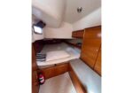 Compact boat cabin with a V-berth bed, wooden storage cabinets, and a small porthole; a fire extinguisher sits on the floor nearby.
