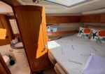 Cozy boat cabin with a large bed, marine-themed cushions, and built-in wooden storage above; a compact head/bathroom is visible nearby.