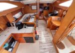 Interior of a wooden sailboat/yacht cabin with a central dining table, L-shaped seating, and a doorway to a forward sleeping cabin.