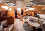 Cozy wooden sailboat interior with a U-shaped dining area, table runner, and cushioned benches; stairs lead to cabins ahead.