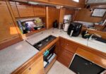 Cozy boat galley with wooden cabinets, stovetop, sink, and counter appliances like a coffee maker and kettle on a speckled countertop.