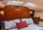 Cozy boat cabin bedroom with wooden walls, two skylights, wall lamps, and a bed with sailboat-themed quilts and a red anchor-pattern pillow.