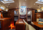 Wood-paneled yacht interior with a curved seating area, table, and doors to cabins; warm lighting and skylights above.