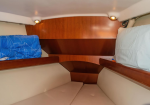 Interior of a small boat cabin with two bunks, beige cushions, and wooden storage panels; a blue blanket on the left bunk and a light on each side.