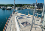 Deck of a sailboat at a sunny marina with hatch covers and rigging, calm blue water, and boats docked nearby.